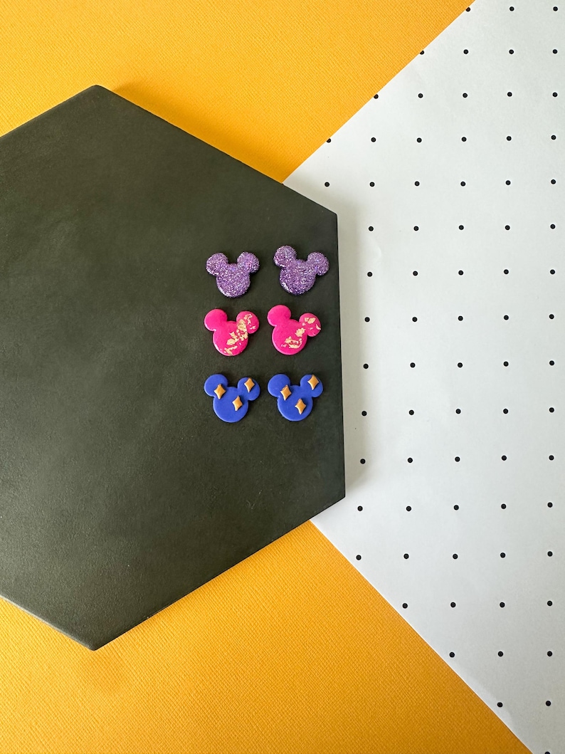 May include: Six colourful Mickey Mouse-shaped stud earrings. Two are purple with glitter, two are pink with gold accents, and two are blue with yellow diamond accents. The earrings are arranged on a dark grey hexagonal surface against a yellow and white background.