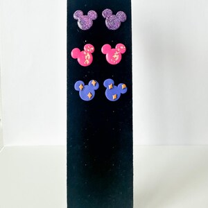May include: A black display stand presents six pairs of stud earrings. The earrings are shaped like Mickey Mouse heads. The top row is glittery purple, the middle row is pink with gold flecks, and the bottom row is blue with gold star accents.