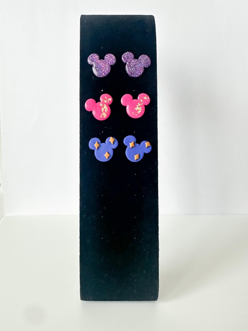 May include: A black display stand presents six pairs of stud earrings. The earrings are shaped like Mickey Mouse heads. The top row is glittery purple, the middle row is pink with gold flecks, and the bottom row is blue with gold star accents.
