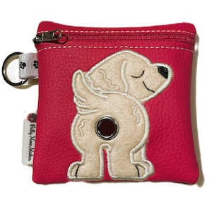 May include: A pink zippered pouch with a white dog embroidered on it. The dog is facing away from the viewer and has its tail curled up. The pouch has a key ring attached to it and says "Billy Now Makes" on the side.