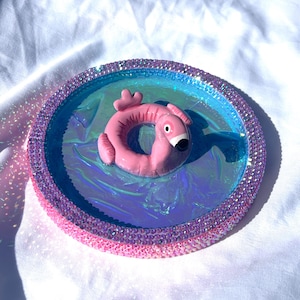 May include: A circular tray featuring a vibrant blue iridescent centre and a rim encrusted with small pink rhinestones. Centred on the tray is a small, three-dimensional pink flamingo-shaped pool float with a black beak and white eye detail.