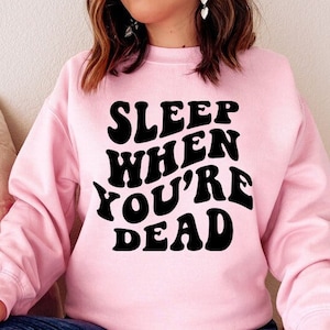 May include: A pink sweatshirt with the text "SLEEP WHEN YOU'RE DEAD" in black retro-style lettering.