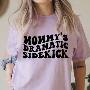 May include: A lavender t-shirt with the text "Mommy's Dramatic Sidekick" in black retro-style lettering.