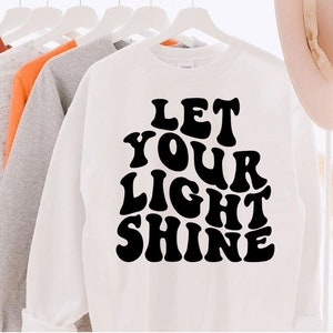 Op de afbeelding: Wit sweatshirt met zwarte tekst die "Let Your Light Shine" luidt in een retro-lettertype.