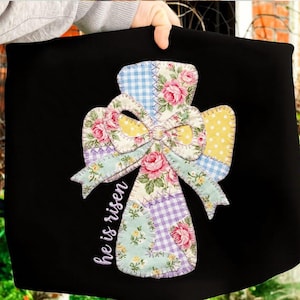 May include: Black fabric with a patchwork cross design. The cross features floral and gingham patterns in pink, blue, yellow, and purple. The words "he is risen" are written vertically in white script.