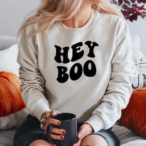 May include: A cream-colored sweatshirt with the words "HEY BOO" in bold, black letters. The sweatshirt is a crew neck style. The person is holding a black mug.