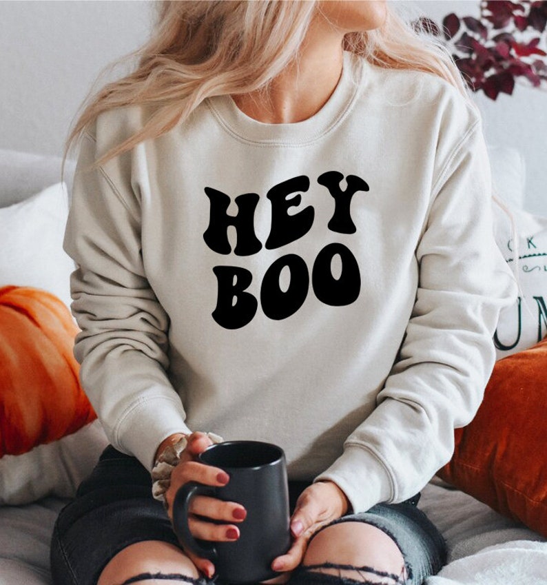 May include: A cream-colored sweatshirt with the words "HEY BOO" in bold, black letters. The sweatshirt is a crew neck style. The person is holding a black mug.