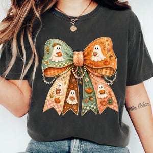 K&ouml;nnte beinhalten: Ein anthrazitfarbenes T-Shirt mit einer gro&szlig;en, dekorativen Schleifengrafik. Die Schleife ist mit kleinen wei&szlig;en Geistern, K&uuml;rbissen, Herbstblumen und karierten Abschnitten in Orange-, Gr&uuml;n- und Cremet&ouml;nen sowie h&auml;ngenden Perlenketten verziert.
