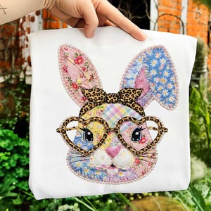 May include: White t-shirt featuring a colorful patchwork bunny face with heart-shaped glasses and a leopard print bow. The bunny's ears are decorated with floral and daisy patterns. The design is outlined with a pearl-like trim.