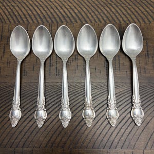 May include: Six silver spoons arranged in a row. Each spoon has a rounded bowl and a decorative handle with ornate detailing. The spoons are set against a dark wood grain surface.