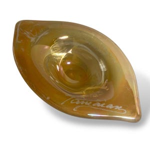 May include: A handcrafted, iridescent glass bowl in a leaf shape. The bowl has a central depression and a wide, shallow rim. The glass has a golden hue with hints of pink and green. The artist's signature, "American", is etched on the side.