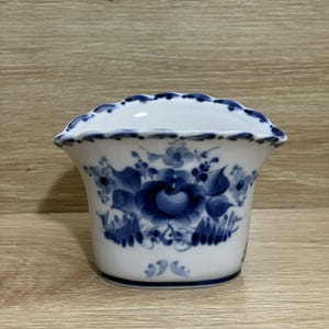 May include: White ceramic vase with a scalloped rim, adorned with blue floral patterns. The vase features a flared shape and a detailed hand-painted design, showcasing a classic, elegant aesthetic. The vase is suitable for displaying flowers or as a decorative piece.