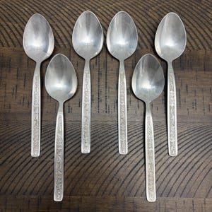 May include: Seven silver spoons with floral etched handles are arranged on a dark wood surface. The spoons have a classic design, with oval bowls and detailed patterns on the handles. The spoons are likely part of a vintage silverware set.