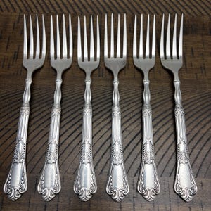 May include: Six silver forks with ornate handles are arranged on a dark wood surface. The forks have tines and decorative detailing on the handles, suggesting a vintage or antique style. The forks are evenly spaced.