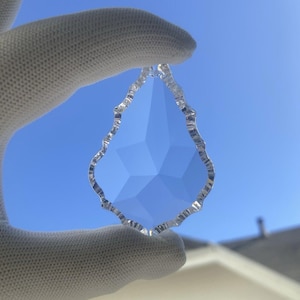 May include: A clear, faceted crystal teardrop pendant with a decorative, scalloped edge. The pendant is held against a bright blue sky, showcasing its geometric interior and reflective qualities. This decorative crystal is ideal for chandeliers or jewelry.