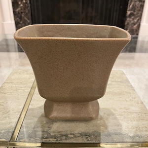 May include: A light brown ceramic planter with a square base and a wide, rounded top. The planter has a speckled texture.