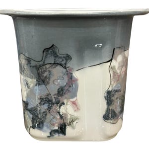 May include: A ceramic vase with a wide rim and a cylindrical body. The upper portion is a muted gray, transitioning to white. Abstract patterns in black, gray, and pink adorn the surface, creating a unique, artistic design.