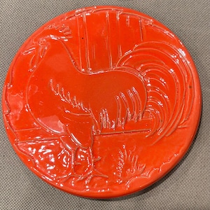 May include: A round, red ceramic coaster featuring a detailed relief of a rooster. The rooster is depicted in profile, with its tail feathers fanned out. The coaster has a glossy finish and is likely used for placing drinks.