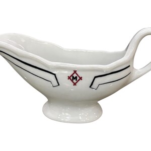 May include: White ceramic gravy boat with a handle and a footed base. The boat features black geometric accents and a red and black emblem. The gravy boat is designed with a pouring spout.