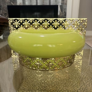 May include: A decorative, lime green glass bowl with a gold-colored, ornate metal base and backing. The bowl has a rounded shape and sits on a clear glass surface. The metal base and backing feature a repeating floral pattern.