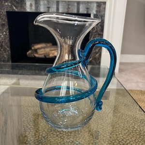 May include: A clear glass pitcher with a turquoise blue handle and a decorative turquoise blue spiral around the body. The pitcher has a flared top and a rounded base. The background includes a fireplace and a glass table.