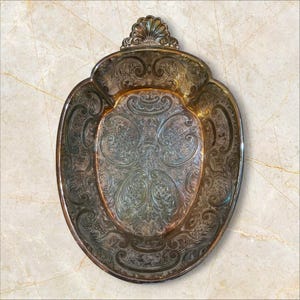 May include: An oval-shaped silver-toned metal tray featuring intricate embossed scrollwork and floral patterns throughout the surface. The tray has a raised scalloped edge at the top and a polished finish with darkened recessed areas to highlight the details.