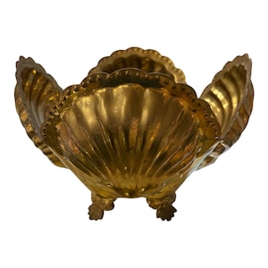 May include: A gold-toned metal bowl shaped like a large scallop shell with a scalloped rim and a textured, ribbed surface. The bowl is supported by three small, decorative feet that are also shaped like miniature shells, providing stability.