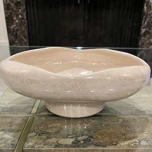May include: A large, pale pink ceramic bowl with a wavy rim and a pedestal base. The bowl has a smooth, glossy finish and a subtle, marbled pattern.