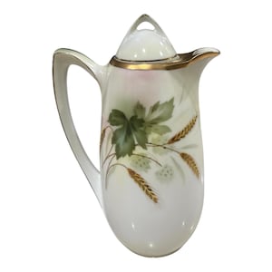 May include: White ceramic pitcher with a lid, handle, and spout. The pitcher is decorated with green leaves, wheat stalks, and gold accents. The rim and handle are trimmed with gold. A decorative piece for serving beverages.