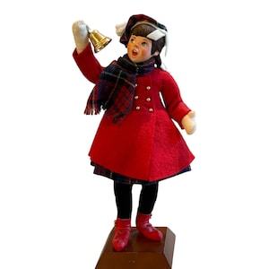 May include: A figurine of a young girl dressed in a red coat, plaid scarf, and hat, holding a gold bell. The figurine has a detailed face and is standing on a wooden base. The girl is wearing black tights and red shoes.