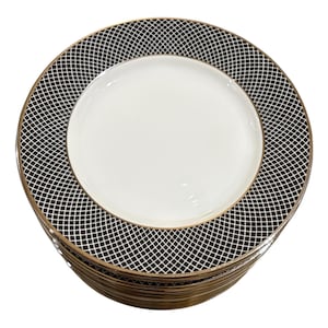 May include: A stack of white ceramic plates with a black and white grid pattern border and a gold rim. The plates have a simple, elegant design, suitable for formal dining or special occasions.