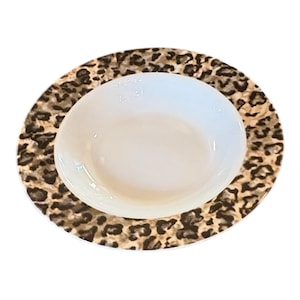 May include: A white ceramic bowl sits atop a leopard print plate. The plate features a brown and black spotted pattern around the rim. The bowl is smooth and glossy, contrasting with the textured plate.