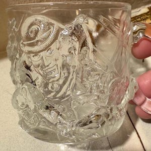 May include: A clear glass mug featuring a raised, embossed design of a smiling figure wearing a bowler hat. The textured surface of the mug displays intricate details of the character's face and clothing, with a sturdy handle on the right side.