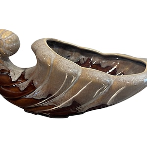May include: A ceramic cornucopia planter with a brown and beige glaze. The planter has a wide, shallow bowl and a curled handle.