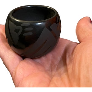 May include: A small, black, hand-painted pottery bowl with a geometric design. The bowl is in the palm of a hand.