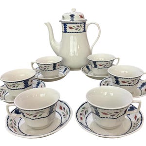 May include: A white porcelain tea set with a teapot and six teacups and saucers. The set features a blue, red, and green leaf design. The teapot has a lid and a handle. The teacups are arranged around the teapot.