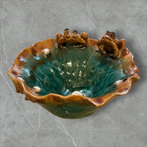 May include: A ceramic bowl shaped like a lotus leaf with a glossy teal interior and brown rim. Two sculpted brown frogs are perched on the scalloped edge, and several small white spherical accents are attached to the inner surface of the bowl.