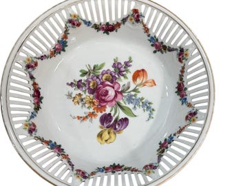 May include: White porcelain serving dish with a pierced rim and a gold edge. The center features a floral bouquet in shades of pink, purple, orange, and yellow. A garland of flowers decorates the rim.