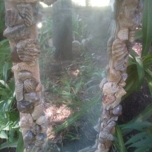 May include: A rectangular mirror with a frame made of various sized stones and shells. The mirror is reflecting a blurry image of trees and foliage.