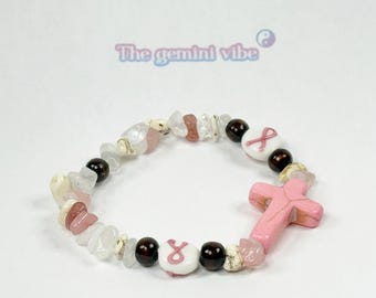May include: A beaded bracelet featuring a pink cross charm, white and pink stone beads, and dark brown beads. The bracelet includes a white bead with a pink ribbon symbol, and the text "The gemini vibe".