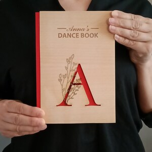 May include: A wooden dance book with a red spine and the text "Anna's Dance Book" on the cover. The cover also features a cutout of the letter "A" with a floral design.