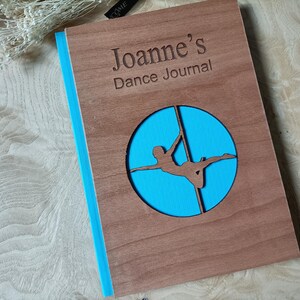 May include: A wooden journal with a blue circle cutout featuring a silhouette of a person on a pole. The journal is personalized with the name "Joanne's Dance Journal".