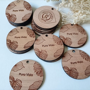 Puede incluir: Colección de etiquetas redondas de madera con acabado natural. Cada etiqueta presenta las palabras "Pura Vida" y un diseño de hojas. Algunas etiquetas tienen un agujero para colgar. Las etiquetas miden unos 5 cm de diámetro.