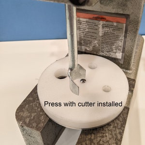 May include: A close-up of a white press with a metal cutter installed. The press has a round base with four holes and the cutter is in the center hole. The text "Press with cutter installed" is written below the press.