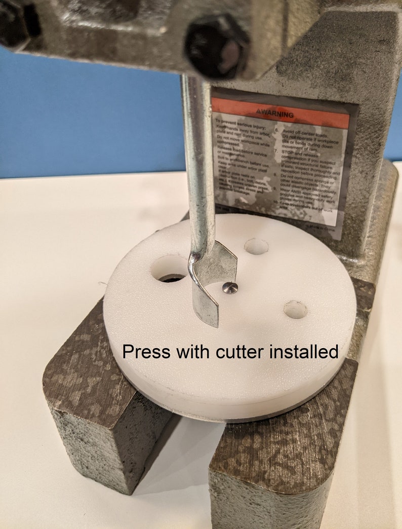 May include: A close-up of a white press with a metal cutter installed. The press has a round base with four holes and the cutter is in the center hole. The text "Press with cutter installed" is written below the press.
