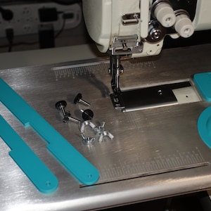 May include: A close-up of a sewing machine with a metal table top. The table top has a ruler, a teal plastic cup, and two teal plastic bars. There are also several metal screws and nuts on the table top.