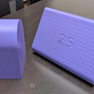 May include: Two light purple objects with a grid pattern embossed on their surfaces. The larger object is a rectangular wedge shape featuring the number 25 centred on its front face, while the smaller object is a tall, narrow, curved block.
