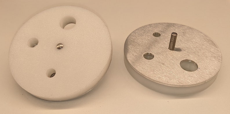 May include: Two round, white and silver metal discs with holes and a metal pin in the center. The discs are likely parts of a machine or device.