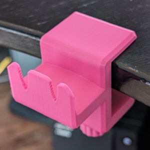 May include: A pink plastic desk organizer with two slots for holding items like pens, pencils, or other office supplies. The organizer is designed to clip onto the edge of a desk or table.