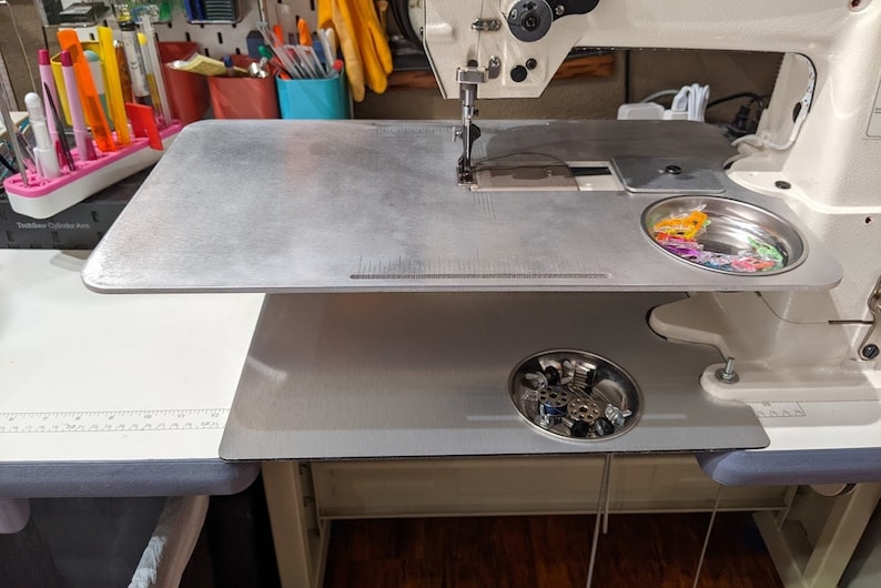 May include: A silver sewing machine table with a ruler and two metal bowls. The top bowl contains colorful plastic bobbins and the bottom bowl contains sewing machine parts.